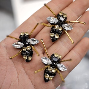 Może przedstawiać: Trzy dekoracyjne broszki w kształcie owadów. Każda broszka ma czarny korpus ozdobiony przezroczystymi i żółtymi kryształami. Nogi owadów to cienkie, złote druciki. Broszki są prezentowane na jasnej powierzchni.