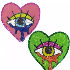 Peut inclure: Deux écussons en forme de cœur ornés de paillettes. L'un est rose avec un motif d'œil bleu, l'autre est vert avec un motif d'œil orange. Les yeux ont des pupilles noires, des paupières jaunes et des larmes de paillettes dégoulinantes.