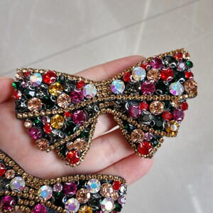 May include: Two bow-shaped embellishments, each adorned with a multitude of colourful rhinestones. The rhinestones are in shades of red, pink, yellow, green, and clear. Gold-coloured beads outline the edges, creating a decorative accessory.