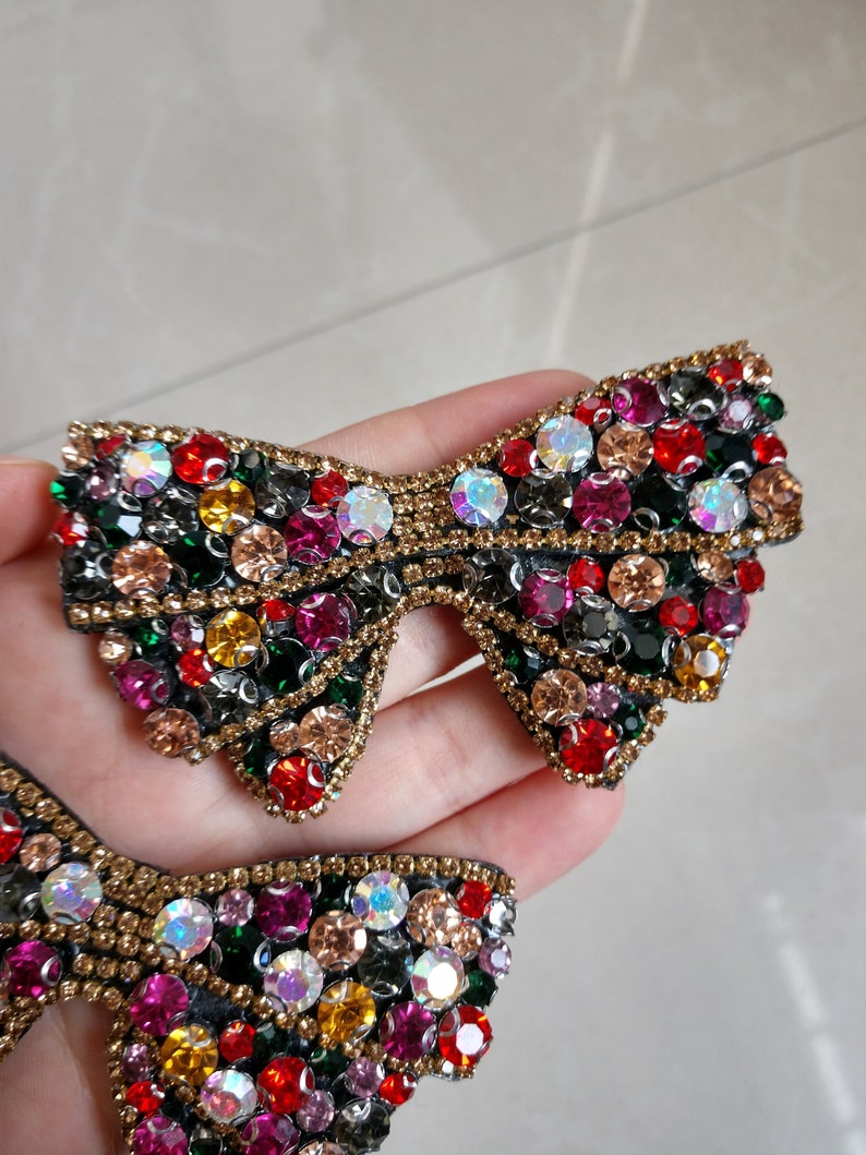 May include: Two bow-shaped embellishments, each adorned with a multitude of colourful rhinestones. The rhinestones are in shades of red, pink, yellow, green, and clear. Gold-coloured beads outline the edges, creating a decorative accessory.