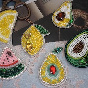 May include: A selection of beaded fruit brooches, including a lemon slice, pear, watermelon slice, and avocado halves. The brooches showcase vibrant colours, detailed beadwork, and gemstone accents. Each brooch is approximately 5-7 cm in size.