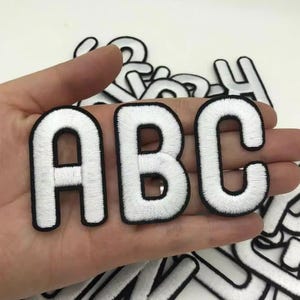 以下が含まれることがあります： 黒い縁取りのある、白い刺繍文字「ABC」3文字。手で持たれており、背景には他の文字パッチが見えます。文字はふっくらとした質感です。