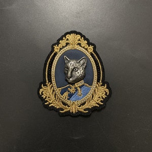 Puede incluir: Un parche bordado en negro y dorado con un leopardo plateado que lleva un uniforme azul y dorado en un marco ovalado dorado.