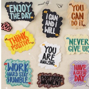 Puede incluir: Diez parches bordados coloridos con citas motivacionales. Los parches presentan frases como "Disfruta del día", "Piensa positivo", "Eres increíble" y "Nunca te rindas".