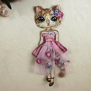 May include: Embroidered iron-on patch of a cat wearing a pink dress with floral details. The cat has brown and white fur, blue eyes, and a pink flower in its hair.