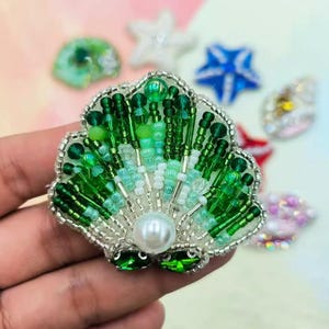May include: A green and white beaded seashell brooch with a pearl center. The brooch is made of glass beads and has a silver backing.