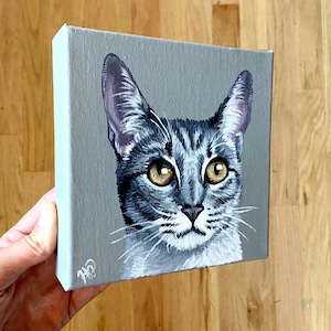 Könnte beinhalten: Ein quadratisches Leinwandgemälde mit einem detaillierten Porträt einer grauen getigerten Katze mit großen bernsteinfarbenen Augen und weißen Schnurrhaaren. Die Katze ist vor einem einfarbigen grauen Hintergrund zentriert, unten links sind Initialen.