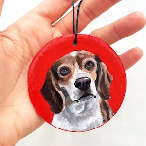 May include: A hand-painted wooden ornament featuring a beagle dog with brown and white fur. The dog is painted on a red background.