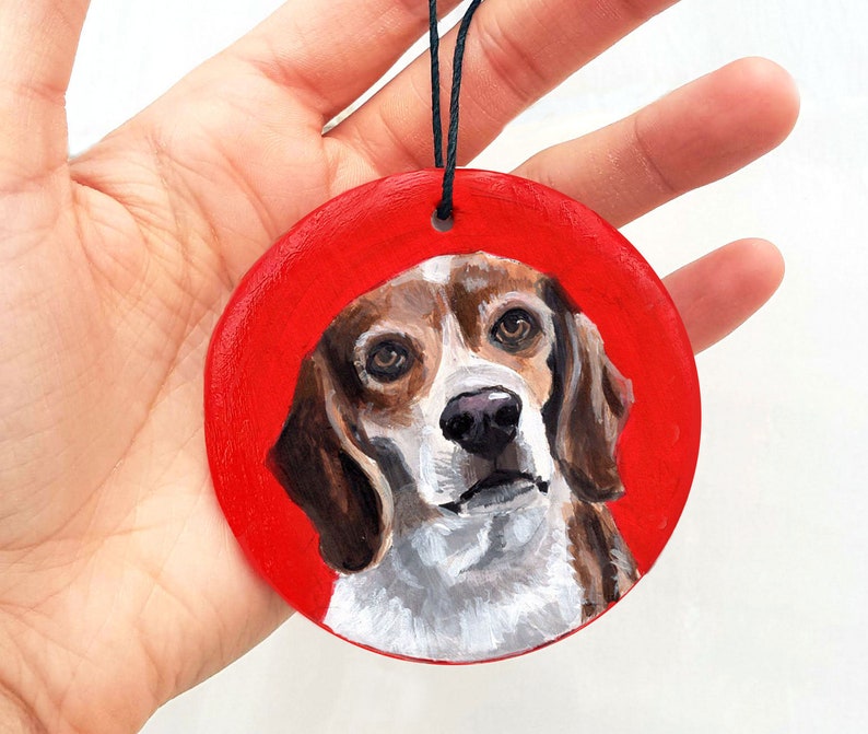 May include: A hand-painted wooden ornament featuring a beagle dog with brown and white fur. The dog is painted on a red background.