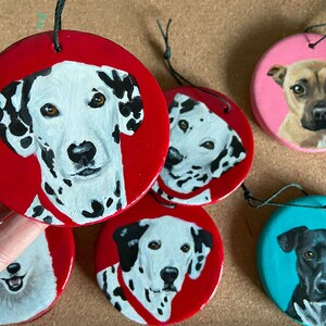 May include: Five hand-painted ceramic ornaments featuring different dog breeds. The ornaments are round and have a string attached for hanging. The dogs are painted in realistic detail and include a Dalmatian, a black and white dog, a brown dog, and a black dog.