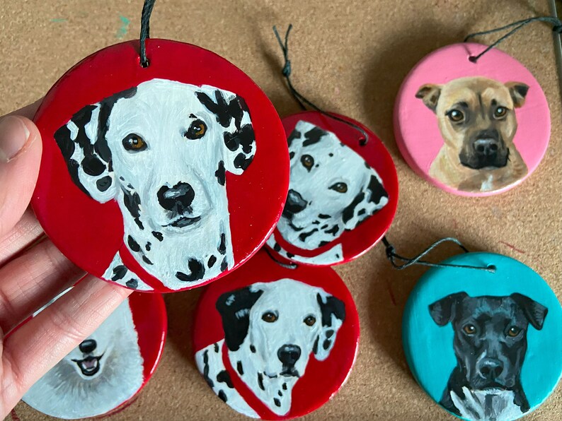 May include: Five hand-painted ceramic ornaments featuring different dog breeds. The ornaments are round and have a string attached for hanging. The dogs are painted in realistic detail and include a Dalmatian, a black and white dog, a brown dog, and a black dog.