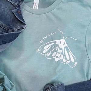 May include: A light blue t-shirt with a white moth graphic and the text "Be the Light" printed on the front.