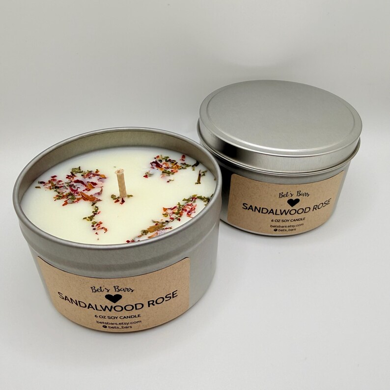 May include: Two silver metal tins of soy candles. One tin is open, revealing a white candle with a wooden wick and dried floral accents. The label reads "Bet's Bars SANDALWOOD ROSE 4 OZ SOY CANDLE". The other tin is closed with the same label.