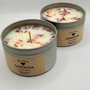 May include: Two round, silver-colored metal tins filled with white wax candles. The candles are decorated with dried petals and have a wooden wick. Each tin has a label that reads "Gardenia" and "6 oz Soy Candle".