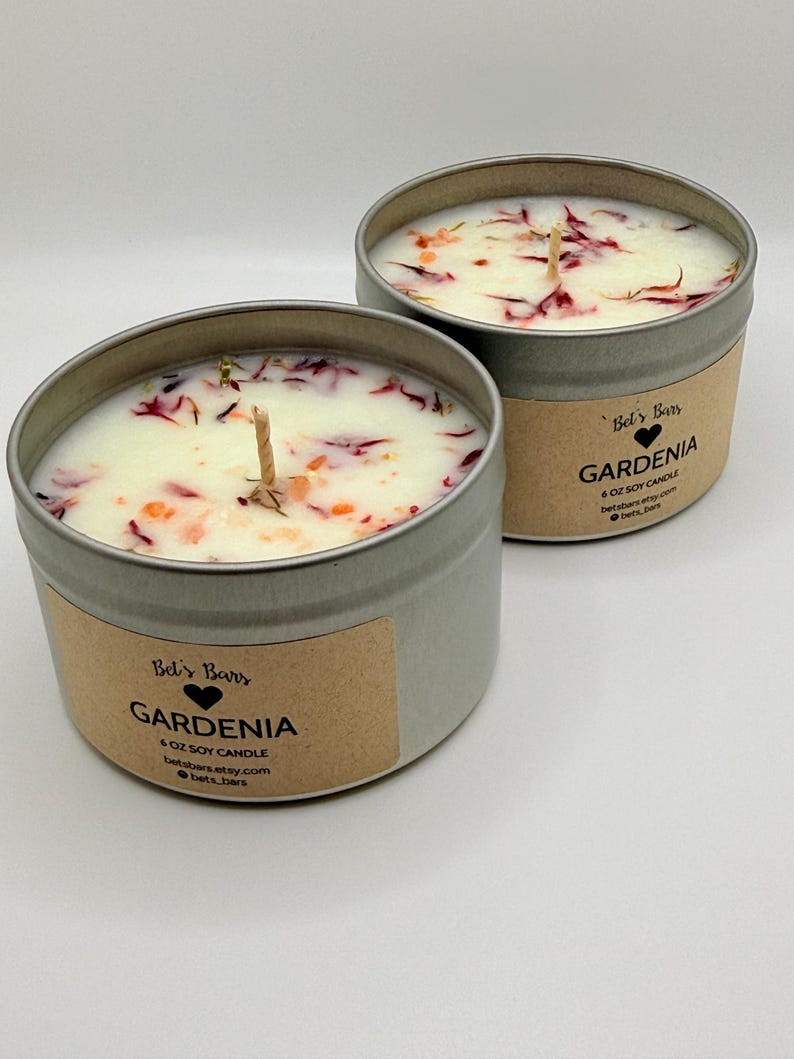 May include: Two round, silver-colored metal tins filled with white wax candles. The candles are decorated with dried petals and have a wooden wick. Each tin has a label that reads "Gardenia" and "6 oz Soy Candle".