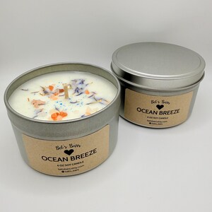 May include: Two silver-colored metal tins of soy candles. The open candle has a white wax base with colorful decorative elements. The label reads "Ocean Breeze" and "6 oz Soy Candle". The second tin is closed with a lid and has the same label.