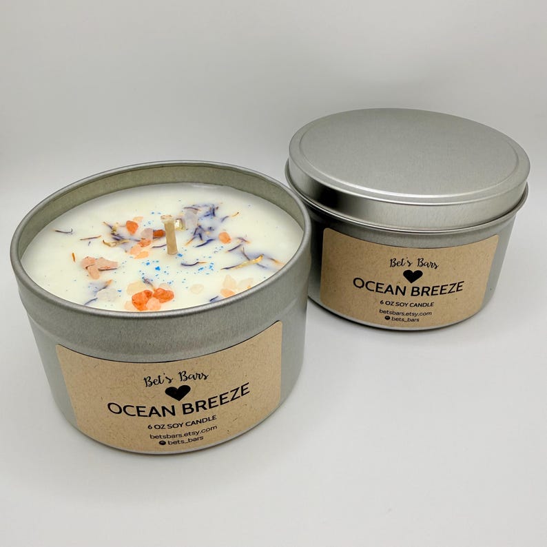 May include: Two silver-colored metal tins of soy candles. The open candle has a white wax base with colorful decorative elements. The label reads "Ocean Breeze" and "6 oz Soy Candle". The second tin is closed with a lid and has the same label.