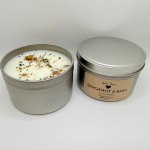 May include: Two silver-colored metal tins, one open and one closed, containing soy candles. The open candle has a white wax with dried herbs and flowers. The closed tin has a label that reads "Bergamot & Basil" and "6 oz Soy Candle".