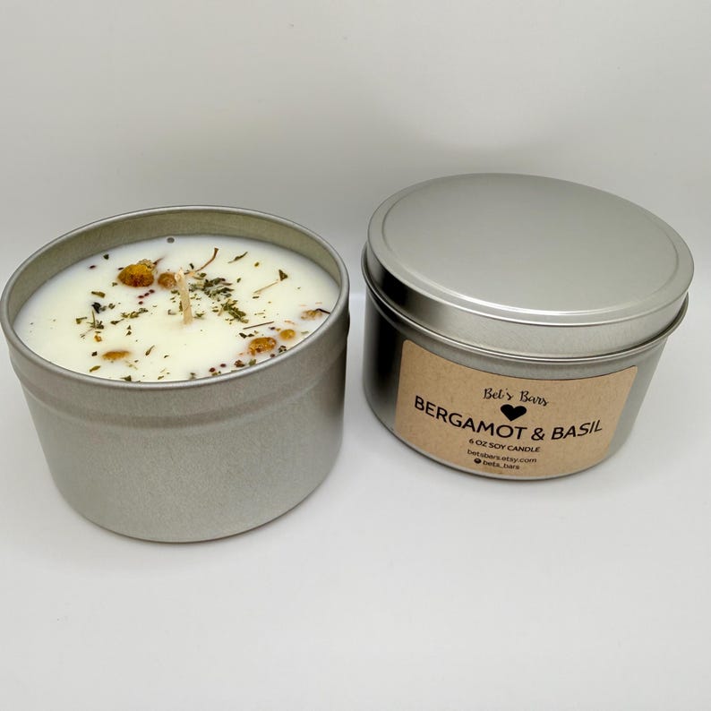 May include: Two silver-colored metal tins, one open and one closed, containing soy candles. The open candle has a white wax with dried herbs and flowers. The closed tin has a label that reads "Bergamot & Basil" and "6 oz Soy Candle".