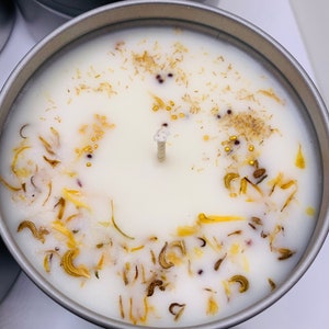May include: Close-up of a white candle in a silver tin, adorned with dried yellow and brown botanicals and small gold beads. The candle has a central wick and is surrounded by other candles.
