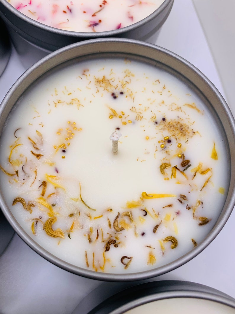 May include: Close-up of a white candle in a silver tin, adorned with dried yellow and brown botanicals and small gold beads. The candle has a central wick and is surrounded by other candles.