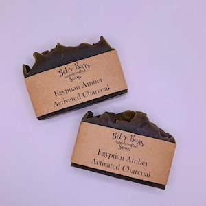 May include: Two bars of dark green soap with brown paper labels. The labels read "Bet's Bars handcrafted soap Egyptian Amber Activated Charcoal".
