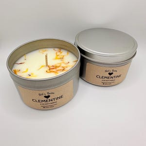 May include: Two 6 oz soy candles in silver tins. One candle is open, revealing a white wax with a wooden wick and orange and yellow botanicals. The label reads "Clementine" and "Bel's Bars". The other candle is closed with a silver lid.