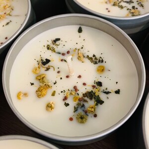 May include: Close-up of several small, round, silver-colored tins filled with white wax candles. Each candle is decorated with dried herbs, seeds, and small yellow flowers. The wicks are visible, suggesting they are ready to be lit.