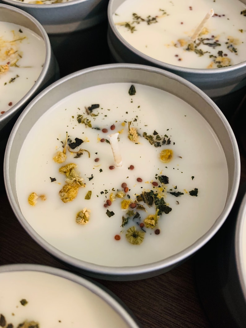 May include: Close-up of several small, round, silver-colored tins filled with white wax candles. Each candle is decorated with dried herbs, seeds, and small yellow flowers. The wicks are visible, suggesting they are ready to be lit.