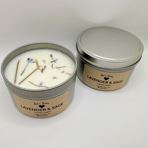 May include: Two silver-colored metal tins of soy candles. One tin is open, revealing a white wax with dried herbs and a wooden wick. The other tin is closed. Both tins have a brown label that reads "Lavender & Sage".