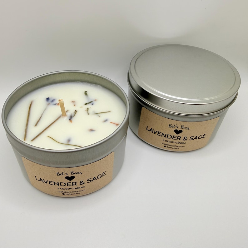 May include: Two silver-colored metal tins of soy candles. One tin is open, revealing a white wax with dried herbs and a wooden wick. The other tin is closed. Both tins have a brown label that reads "Lavender & Sage".