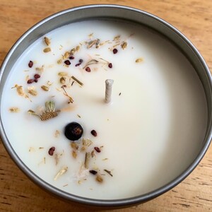 May include: A round, silver-colored tin candle filled with white wax. The candle is decorated with dried herbs, seeds, and a dark berry. A single, unlit wick is centered in the wax.
