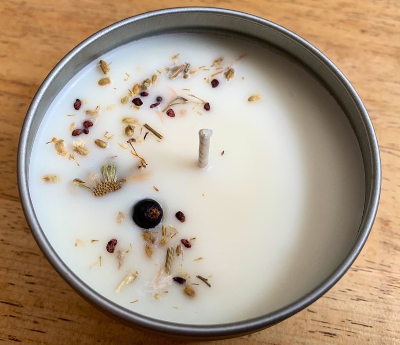 May include: A round, silver-colored tin candle filled with white wax. The candle is decorated with dried herbs, seeds, and a dark berry. A single, unlit wick is centered in the wax.