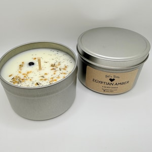 May include: Two silver metal tins, one open and one closed, containing soy candles. The open tin reveals a white candle with a wooden wick and decorative herbs. The closed tin has a label that reads "Egyptian Amber" and "Bel's Bars".