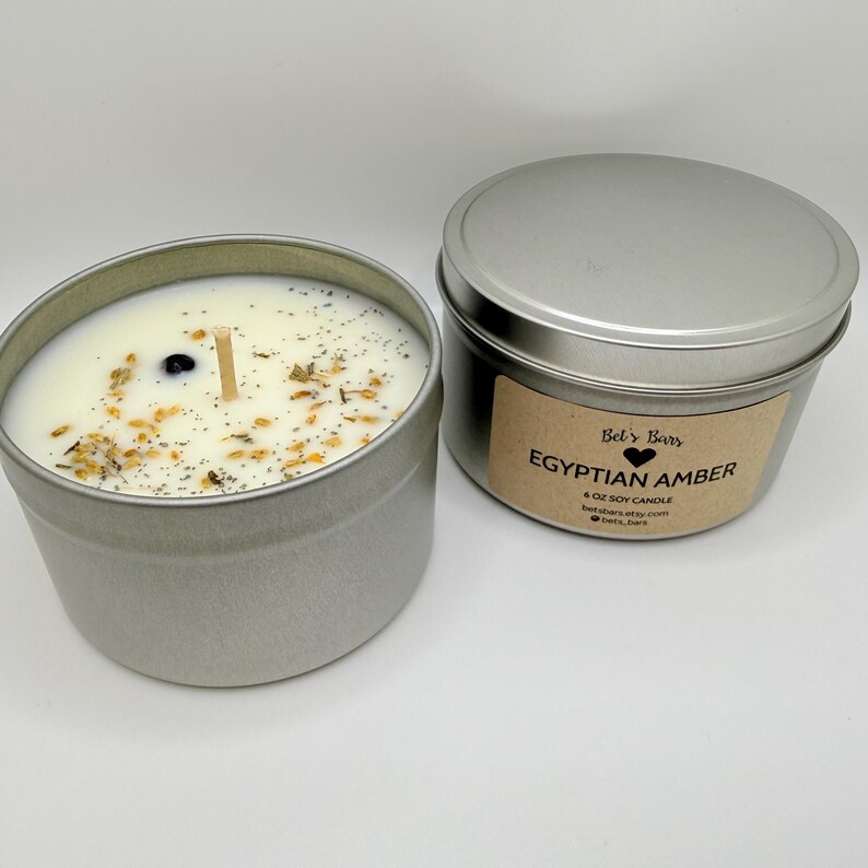 May include: Two silver metal tins, one open and one closed, containing soy candles. The open tin reveals a white candle with a wooden wick and decorative herbs. The closed tin has a label that reads "Egyptian Amber" and "Bel's Bars".