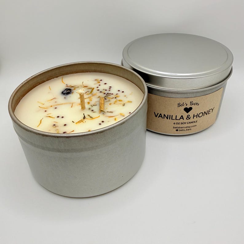 May include: A soy candle in a silver tin with the text "Vanilla & Honey" on the label. The candle wax is off-white and decorated with dried botanicals. The candle is 6 oz.
