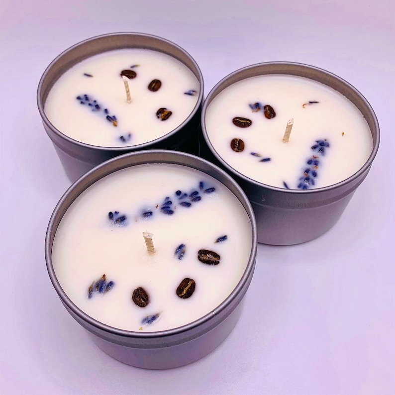 May include: Three small, silver-colored tin candles with white wax, each decorated with coffee beans and lavender sprigs. The candles have wooden wicks. The background is a solid white.