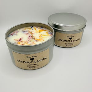 May include: Two silver-colored metal tins of Coconut & Santal scented soy candles. The candles have a white wax base with colorful dried flower petals. Each tin has a brown label with the text "Coconut & Santal" and the brand name "Bet's Bars".