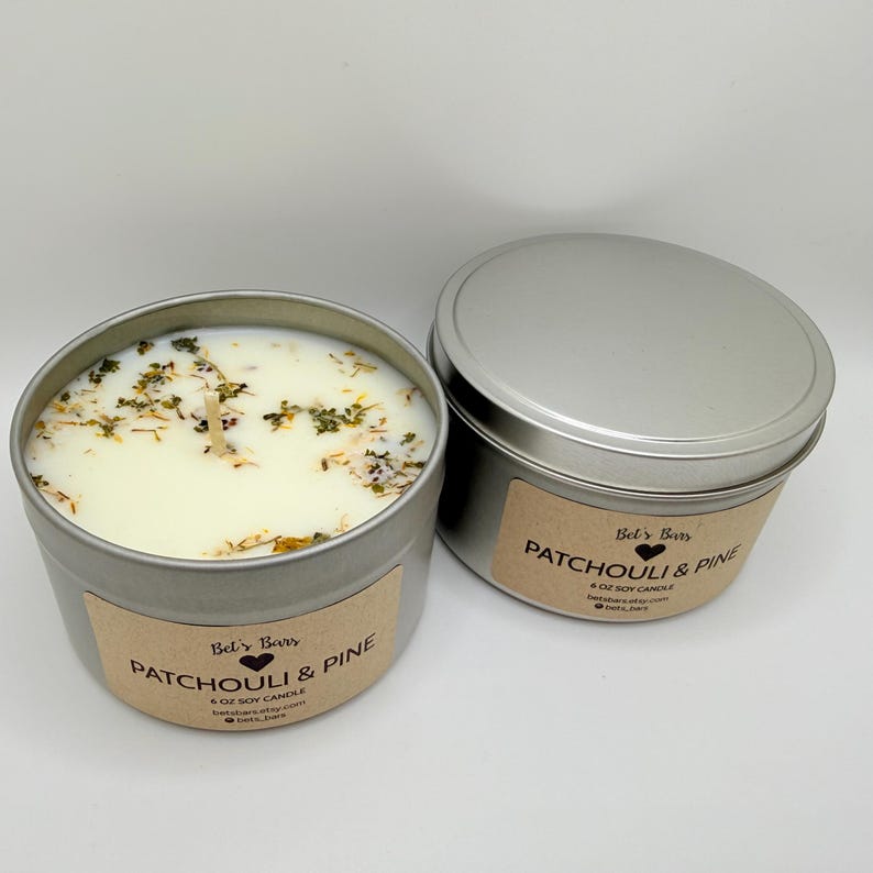 May include: Two silver-colored, round soy candles with lids. One candle is open, revealing a creamy wax with dried herbs. The labels read "Patchouli & Pine" and "Bel's Bars". The open candle has a wooden wick.