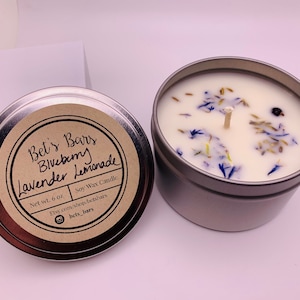 May include: A silver-colored tin candle with a white wax interior, adorned with dried blue flowers and a central wick. The label reads "Bet's Bars Blueberry Lavender Lemonade" and indicates a net weight of 6 oz.