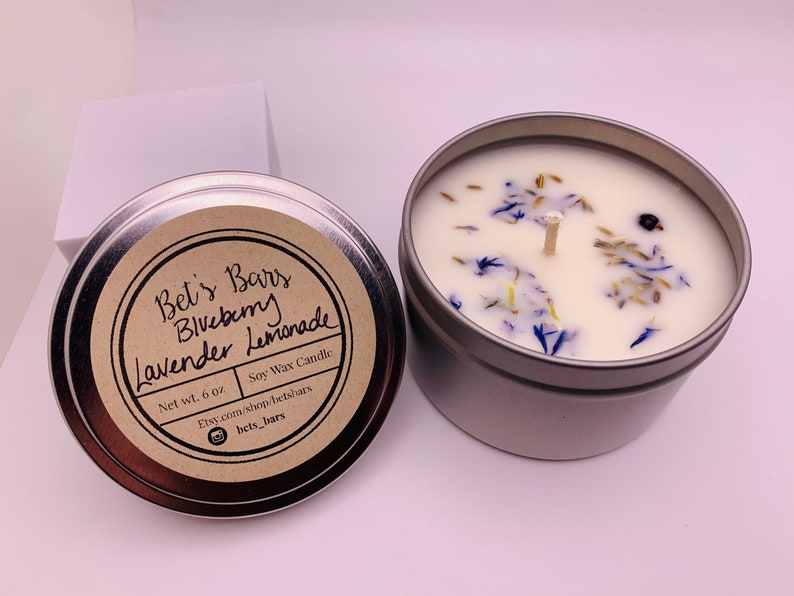 May include: A silver-colored tin candle with a white wax interior, adorned with dried blue flowers and a central wick. The label reads "Bet's Bars Blueberry Lavender Lemonade" and indicates a net weight of 6 oz.