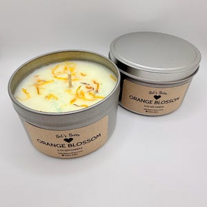 May include: Two metal tin candles with lids. The candles are labeled "Orange Blossom" and contain a light-colored wax with dried orange petals. The labels also include the text "6 OZ SOY CANDLE" and the brand name "Bet's Bars".
