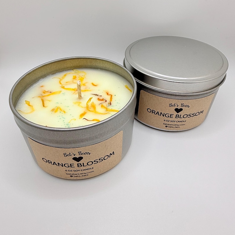 May include: Two metal tin candles with lids. The candles are labeled "Orange Blossom" and contain a light-colored wax with dried orange petals. The labels also include the text "6 OZ SOY CANDLE" and the brand name "Bet's Bars".