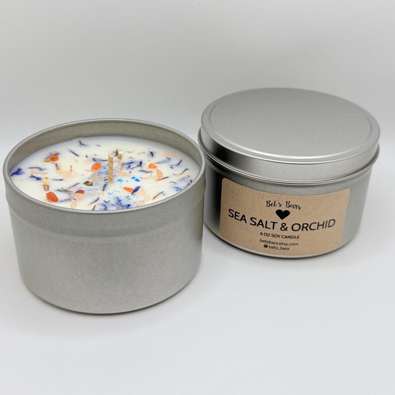 May include: Two silver-colored, round metal containers. One is open, revealing a white candle with a wooden wick and colorful floral accents. The other is closed, with a label that reads "Sea Salt & Orchid" and "6 OZ SOY CANDLE".