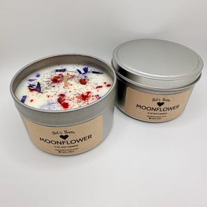 May include: Two silver-colored metal tins, one open and one closed, each containing a candle. The open candle has a white wax base with red, blue, and green decorative elements. The label reads "Moonflower" and "6 oz Soy Candle".