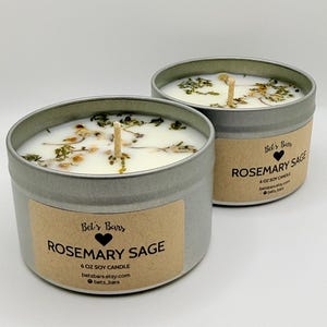 May include: Two Rosemary Sage soy candles in silver tins. Each candle has a wooden wick and is adorned with dried herbs. The labels read "Rosemary Sage" and "6 oz Soy Candle". The brand name "Bel's Bars" is also visible.