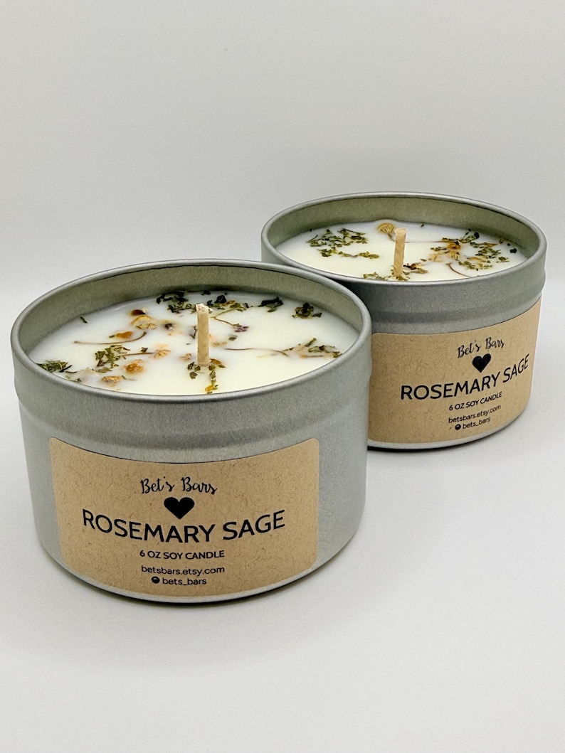May include: Two Rosemary Sage soy candles in silver tins. Each candle has a wooden wick and is adorned with dried herbs. The labels read "Rosemary Sage" and "6 oz Soy Candle". The brand name "Bel's Bars" is also visible.
