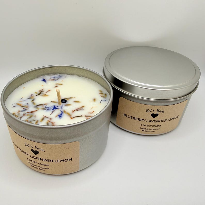May include: Two silver-colored tin candles with lids. One candle is open, revealing a white wax with dried flowers and a wick. The label reads "Blueberry Lavender Lemon" and "6 oz Soy Candle". The other candle is closed.