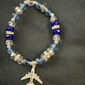 Puede incluir: Una pulsera de cuentas con un dije de avión plateado. La pulsera tiene cuentas transparentes, azules y azul oscuro, con detalles plateados. El dije de avión está detallado con pequeños adornos.