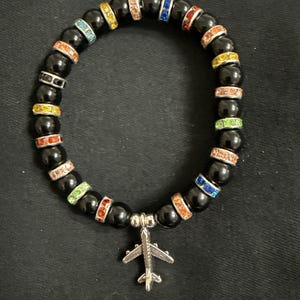 May include: Black beaded bracelet with colourful rhinestone accents and a silver airplane charm. The bracelet features black beads interspersed with bands of rhinestones in various colours, including red, orange, yellow, green, and blue. The airplane charm hangs from the centre.